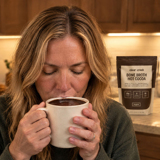 A woman sipping a mug of thick Clear Creek bone broth hot cocoa in her kitchen, the pouch on the counter behind her
